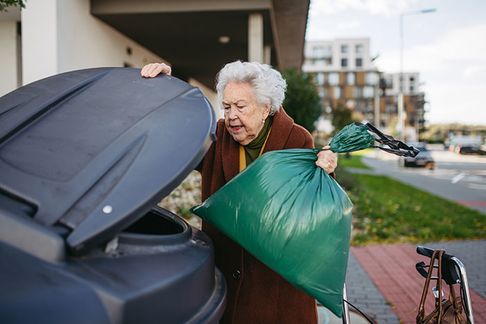 Elderly woman digging through trash with a green bag, illustrating MIL banned from visiting couple after secret digging discovery.