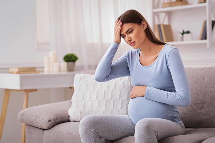 Pregnant woman sitting on couch holding stomach and forehead, portraying stress in a home setting.