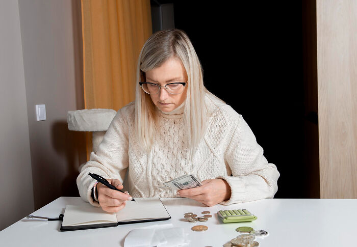 Woman calculating finances at table, holding cash and writing in notebook, illustrating hidden huge debt situation.