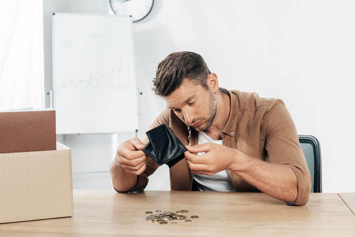 Man sitting at table, looking distressed while checking empty wallet and counting few coins, facing huge debt issue.