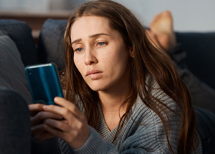Woman looking suspicious and confused while checking phone during husband's surgery at home on the couch.