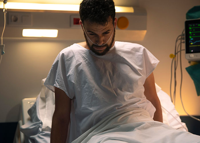 Man in hospital gown sitting on bed, appearing worried during surgery, reflecting suspicion and confusion of wife checking phone.