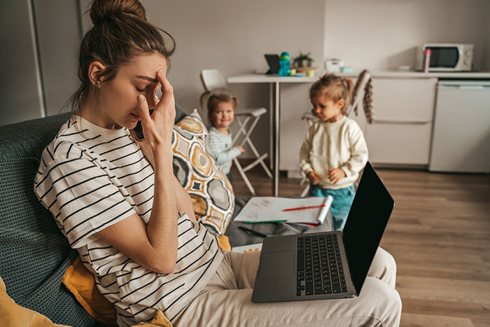 Stressed woman working on laptop at home while managing full-time work, study, and caring for two kids.