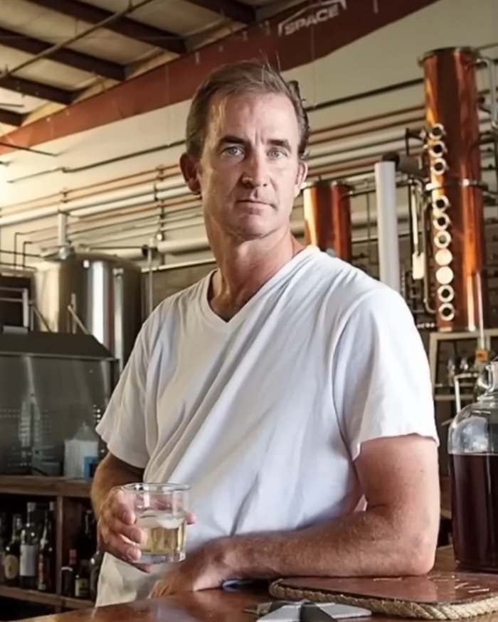 Man in white shirt holding a drink inside an industrial setting linked to Coldplay KissCam saga and HR exec concert twist.