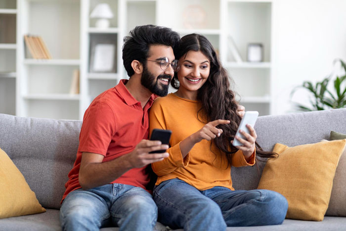 Couple sitting on a couch smiling and looking at phones, discussing husband parents native language at home.