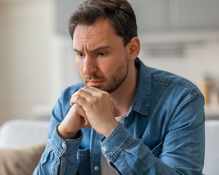 Worried husband sitting indoors, looking stressed and deep in thought while new mom struggles with baby care.