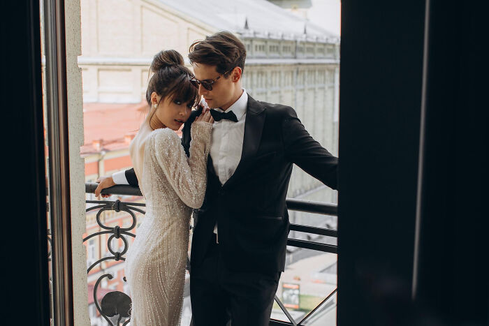 Woman in a white dress and man in a black tuxedo posing closely on a balcony, illustrating reasons women get the ick from a guy.