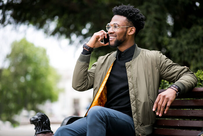 Man sitting on park bench, smiling and talking on phone, illustrating moments when women get the ick from a guy.
