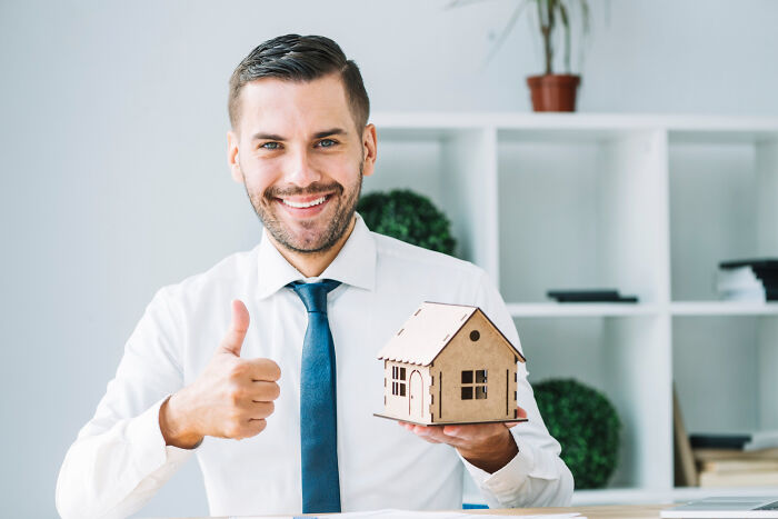 Smiling man in a white shirt and blue tie holding a small wooden house model, giving a thumbs up gesture.