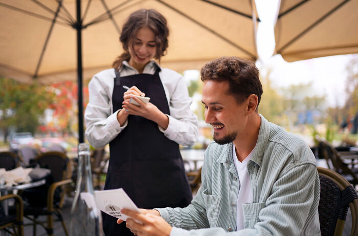 Man smiling at menu while waitress takes order outdoors, capturing a moment that could cause the ick from a guy.