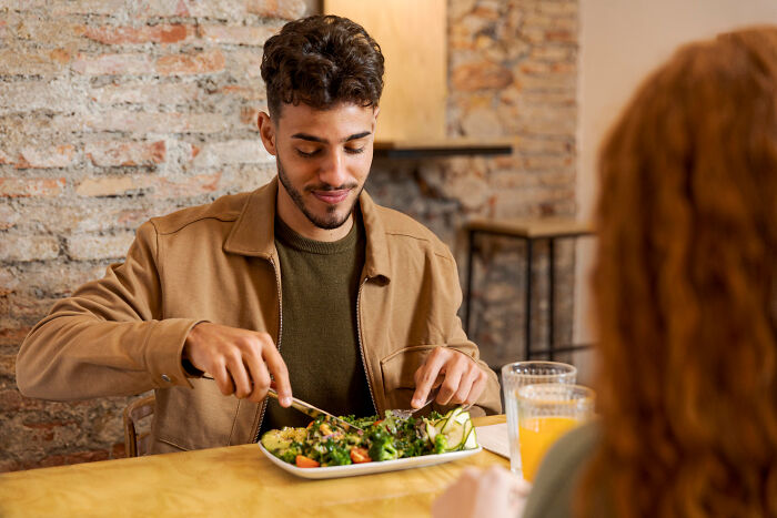 Young man looking hesitant eating salad on a date while woman watches, capturing reasons why women get the ick from a guy