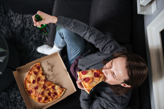Woman sitting on couch eating pizza and holding a beer bottle, illustrating reasons women get the ick from a guy.
