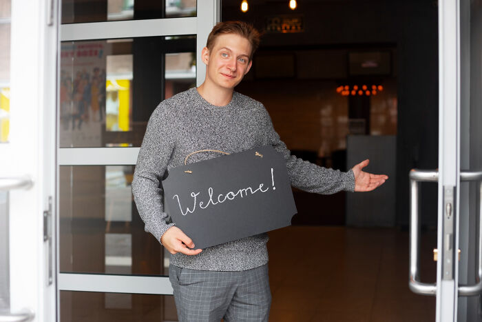 Young man holding welcome sign and gesturing inside a building, related to reasons women got the ick from a guy.