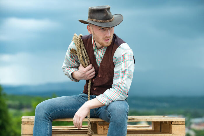Man in cowboy hat and vest sitting on wooden pallets holding rope, illustrating reasons women got the ick from a guy.