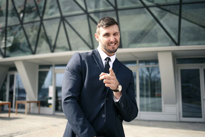 Man in a suit winking and pointing outside a modern building, capturing reasons why women got the ick from a guy.