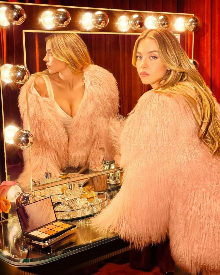 Sydney Sweeney in a pink fur coat at a vanity mirror surrounded by makeup, highlighting business ventures and net worth. Sydney Sweeney in a pink fur coat at a vanity mirror surrounded by makeup, highlighting business ventures and net worth.