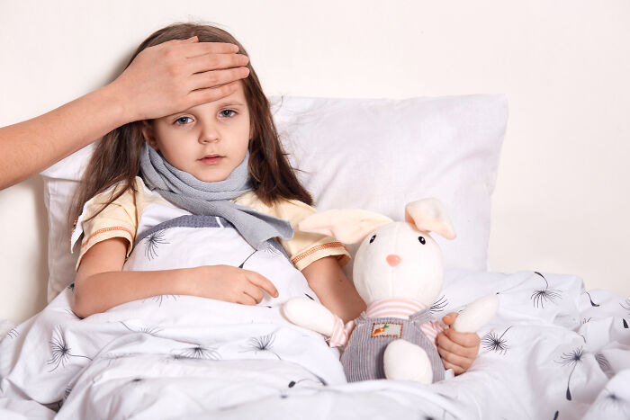 Sick young girl in bed with scarf holding stuffed bunny while an adult checks her forehead, illustrating loss of savings event.