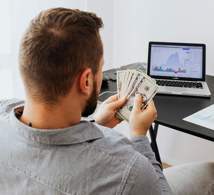 Man counting cash while reviewing financial charts on laptop, illustrating earning more than girlfriend concept.