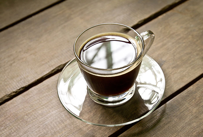 Clear glass cup filled with black coffee on a glass saucer placed on a wooden table for daily dose knowledge.