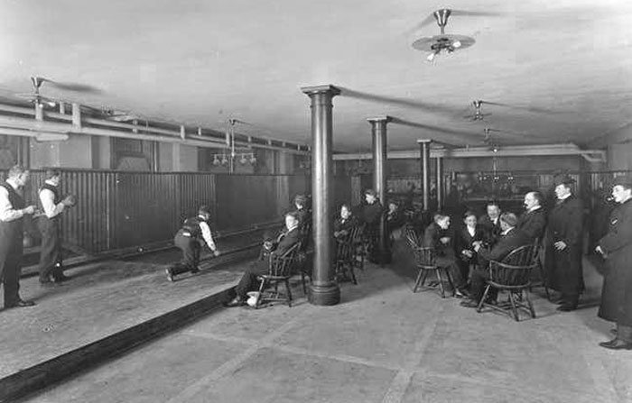 Group of men in early 20th-century attire playing and watching indoor bowling in a vintage recreation room, historical knowledge fact.