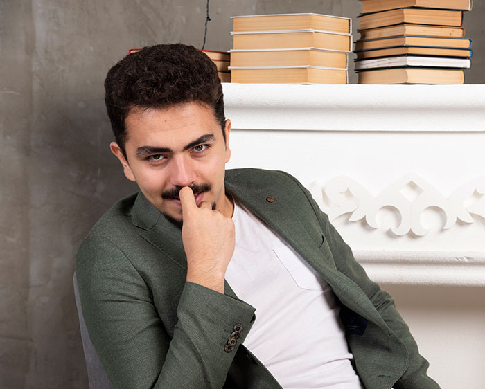 Young man in green blazer posing thoughtfully near stacked books, representing daily dose of fascinating knowledge facts