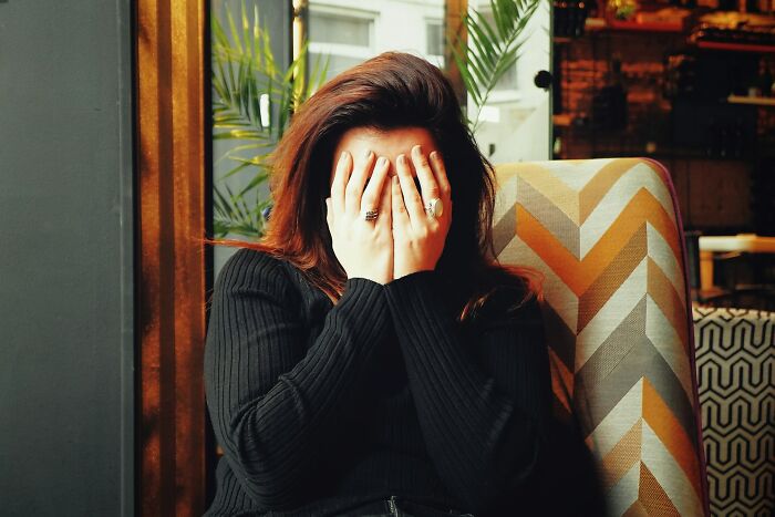 Person in a black sweater covering their face with hands while sitting on a patterned chair indoors, expressing emotion.