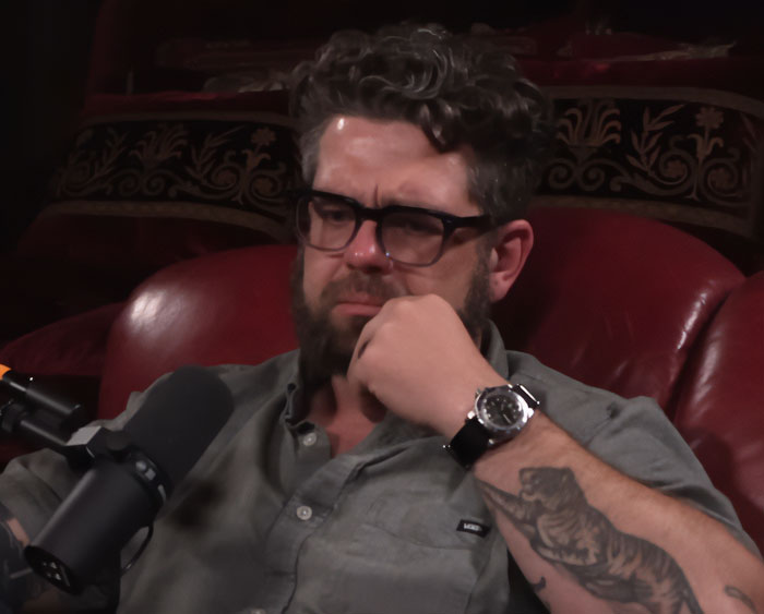 Man with glasses and a beard wearing a gray shirt, deep in thought during a podcast on idiocy and nonsense debates. Man with glasses and a beard wearing a gray shirt, deep in thought during a podcast on idiocy and nonsense debates.
