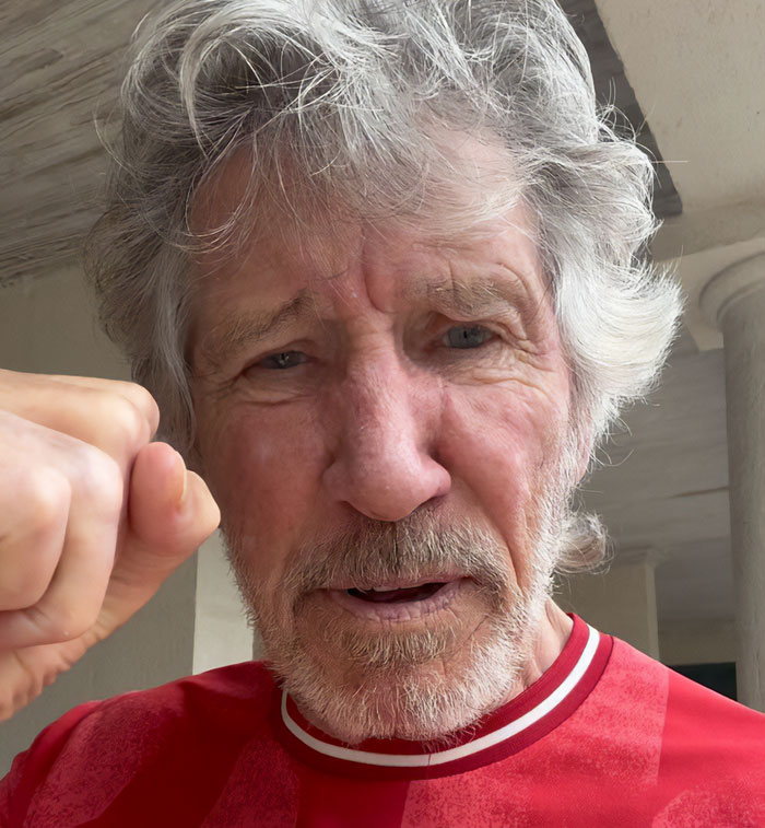 Roger Waters with a raised fist, expressing anger and intensity in a casual indoor setting. Roger Waters with a raised fist, expressing anger and intensity in a casual indoor setting.
