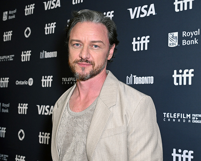 James McAvoy posing at Toronto Film Festival event, looking at camera in beige jacket and gray shirt.