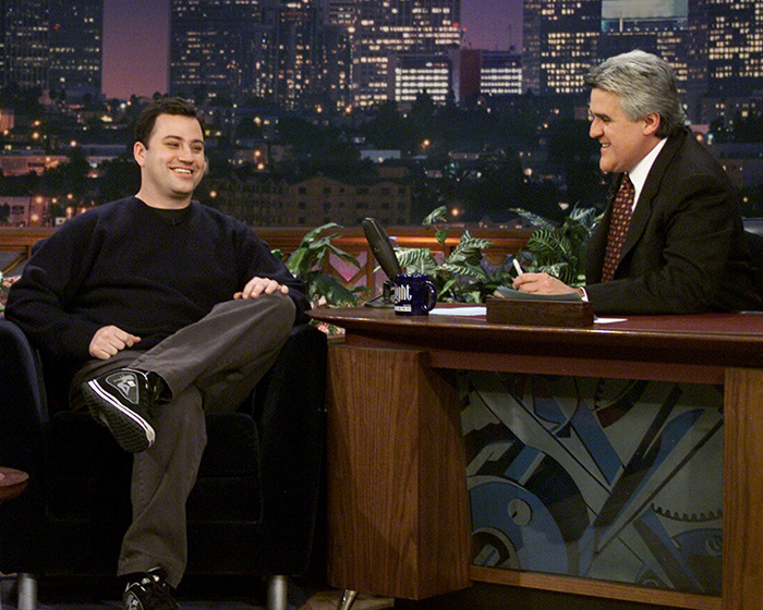 Jay Leno hosting a late-night talk show, engaging in conversation with a seated guest in a studio setting.