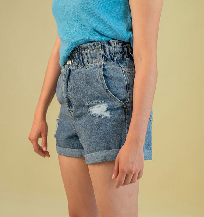 Woman wearing distressed mom shorts and a blue top standing against a plain neutral background.