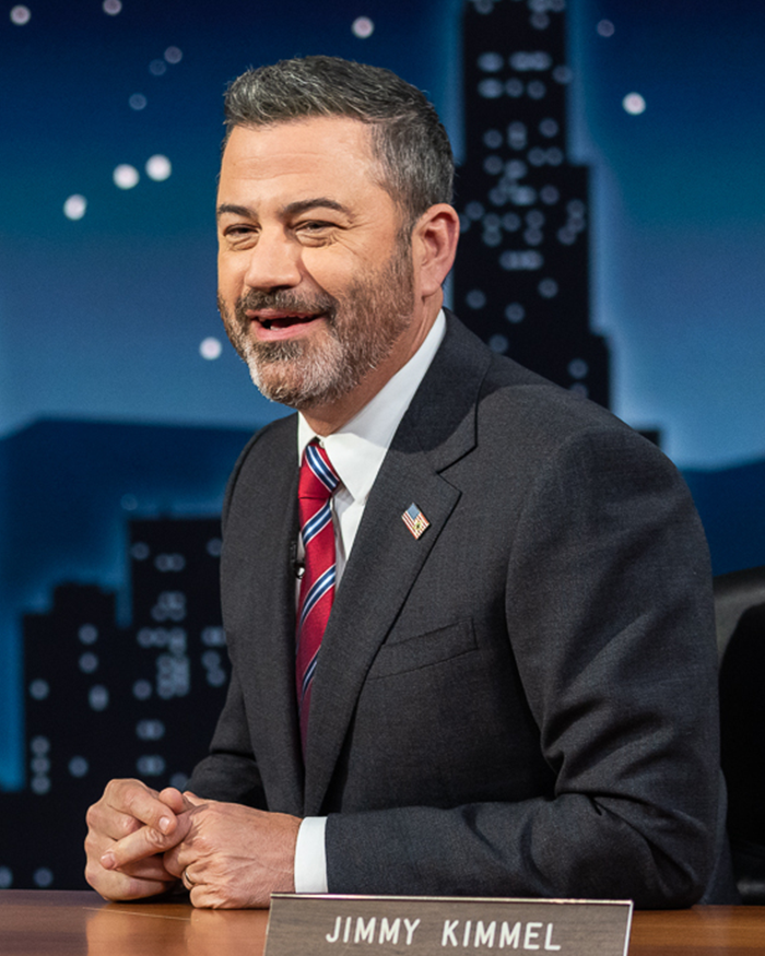 Jimmy Kimmel smiling in a dark suit and striped tie on a late night show set with a city skyline background.
