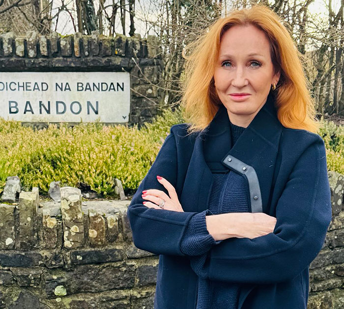Red-haired woman standing outdoors near a stone wall and sign, representing J.K. Rowling team addressing viral cancer rumor. Red-haired woman standing outdoors near a stone wall and sign, representing J.K. Rowling team addressing viral cancer rumor.