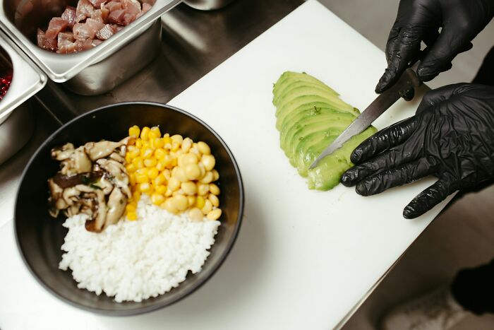 Person wearing black gloves slicing avocado next to a bowl of rice, corn, and mushrooms in a restaurant kitchen health violation context