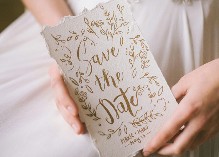 Bride in a white wedding dress holding a floral save the date card with gold calligraphy text.
