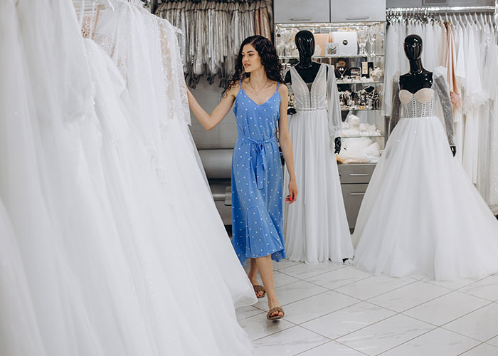 Woman in a blue dress browsing white wedding dress options in a bridal boutique with mannequins displaying gowns nearby.