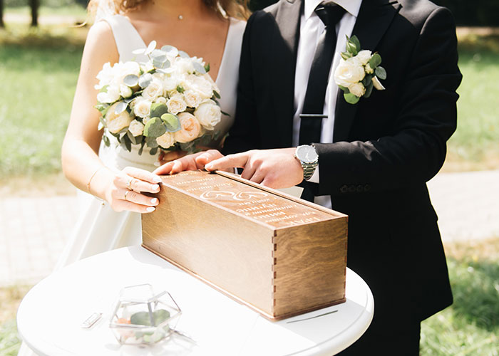 Bride in a white wedding dress holding a bouquet with groom in suit opening a wooden box outdoors at a wedding ceremony.