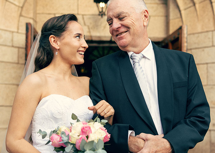 Bride in a white wedding dress holding a bouquet, smiling at an older man in a dark suit outside a stone building.