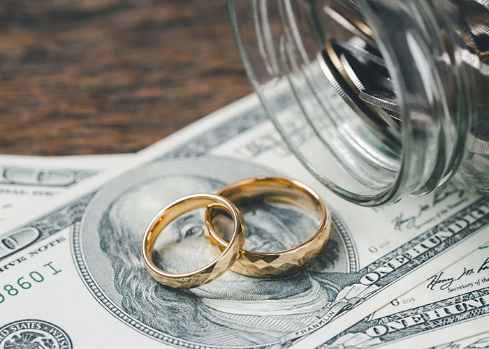 Two gold wedding rings resting on a hundred-dollar bill next to a tilted jar of coins, symbolizing wedding expenses and white wedding dress.