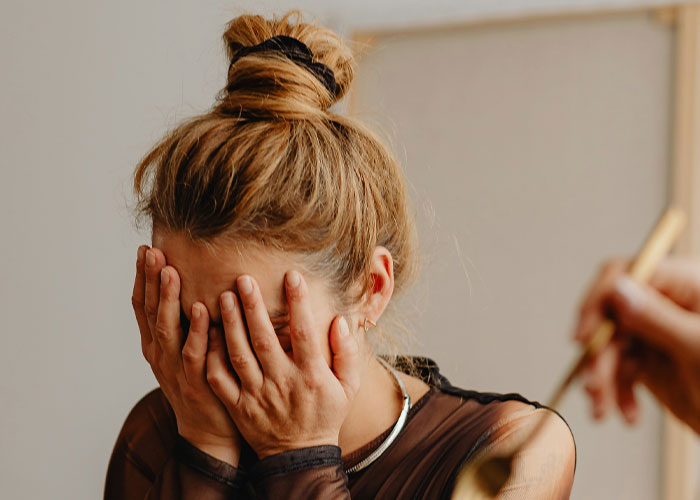 Woman with blonde hair in a messy bun covering her face with hands, wearing a black top in a neutral indoor setting.