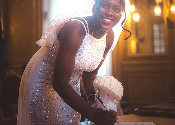 Bride in sparkling white wedding dress signing a document indoors, holding a white floral bouquet and smiling warmly.