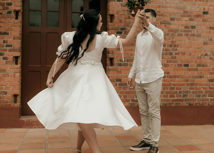 Bride in a flowing white wedding dress twirling while holding hands with groom outside a brick building during wedding photoshoot.