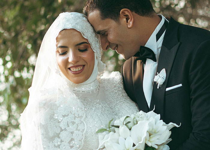 Bride wearing a white wedding dress with intricate lace details posing with groom in a black tuxedo holding white flowers.