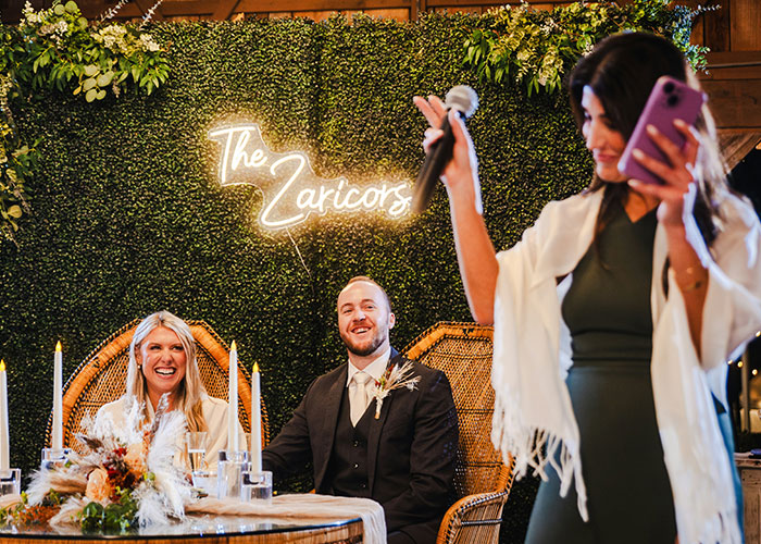 Bride in a white wedding dress and groom seated at a decorated table during a wedding speech with greenery backdrop.