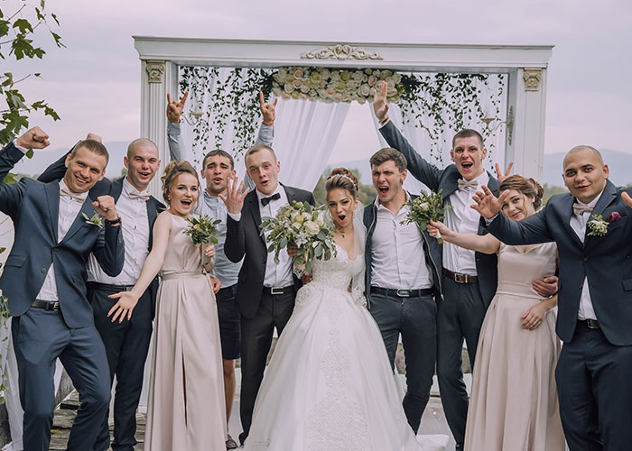 Bride in a white wedding dress and groom celebrate with their bridal party under a floral wedding arch outdoors.