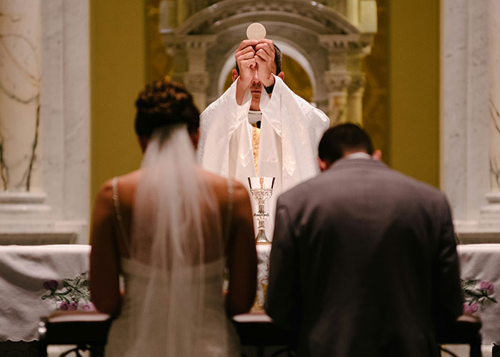 Bride in a white wedding dress and groom kneeling at the altar during a traditional wedding ceremony with a priest.