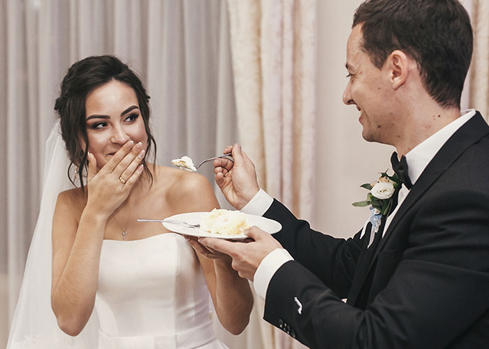 Bride in a white wedding dress smiling shyly as groom feeds her a piece of wedding cake during reception celebration.