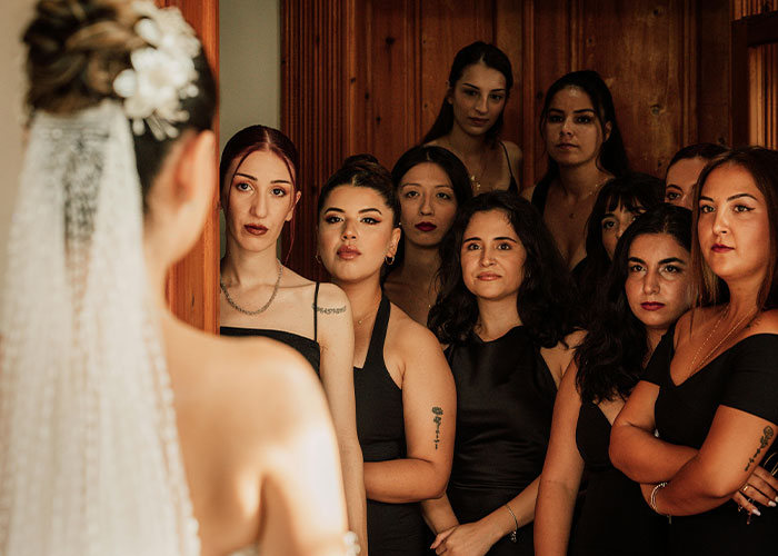 Bride in a white wedding dress with veil, surrounded by bridesmaids in black dresses, preparing indoors before ceremony.