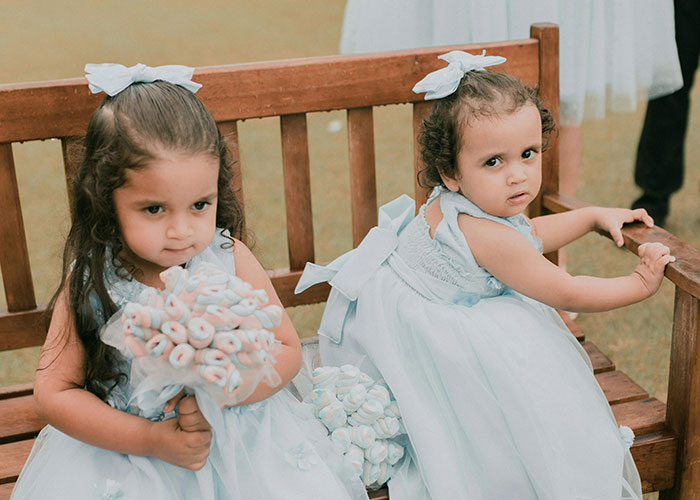 Two young girls in white wedding dresses sitting on a wooden bench, one holding a bouquet, outdoors at a wedding.