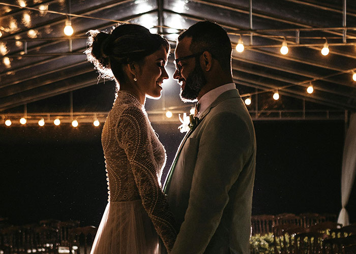 Bride in a white wedding dress holding hands with groom under string lights during evening wedding celebration.